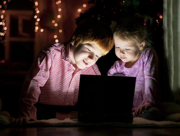 children sitting on floor in dark room playing with laptop on background of glowing garland lights