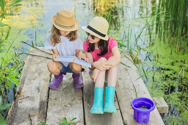 自然の中でノートを読んで遊んで休んでいる2人の女の子の子供たち。木製の湖の桟橋に座っている子供たち、夏の日没の水の風景の背景、カントリースタイル