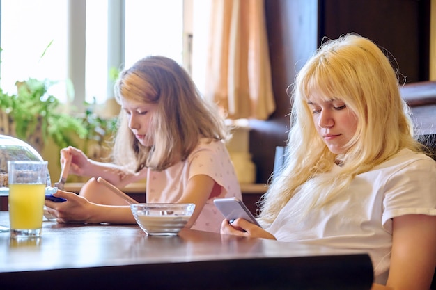 Children two girls sisters eating at home looking at smartphones