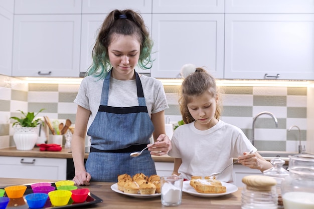 Children two girls sisters preparing muffins in home kitchen. Kids sprinkling decorating freshly baked homemade cakes with white sugar powder