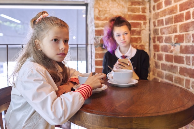 Children, two sisters, teenager and younger sister, sitting together in cafe at table. Girls resting after school