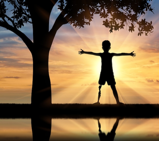 Children with disabilities concept. Happy disabled boy with a prosthetic leg standing near a tree at sunset and the river with its reflection