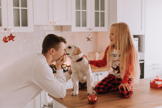 愛犬を連れた子供たちがクリスマスキッチンに座っています。家族の娯楽。ライフスタイル。兄と妹、男の子と女の子はペットと遊んだり、だましたりします。テキスト用のスペース。高品質の写真