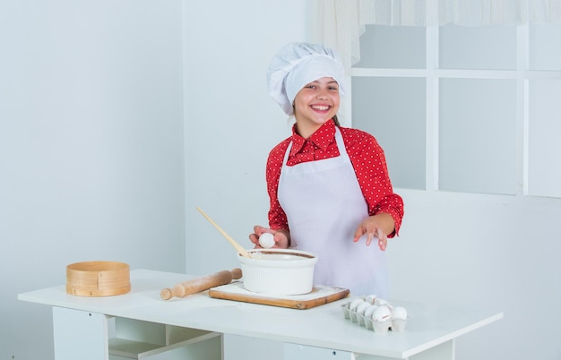 子供の日はキッチンでクッキーを焼くプロの熟練したパン屋の子供シェフの制服と帽子の十代の女の子がレシピの時間でケーキを作る生地を準備してキッチンで幸せな子供料理を食べる