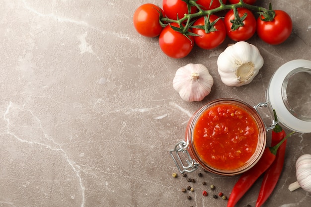 Salsa di peperoncino rosso in barattolo di vetro ed ingredienti su fondo grigio, spazio per testo