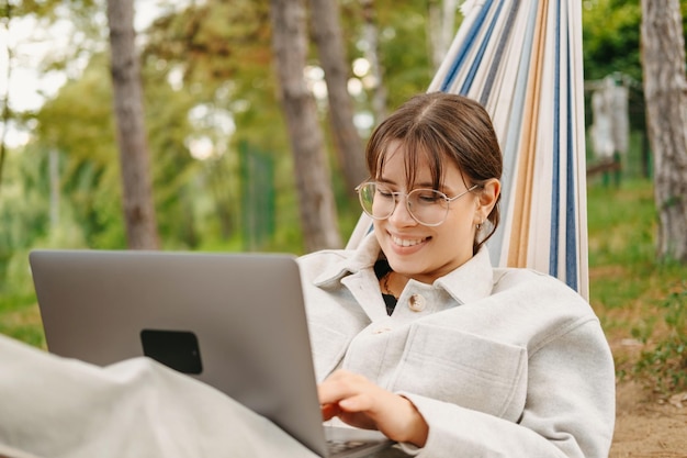 Chill lachende jonge vrouw zit in een hangmat en werkt op de laptop