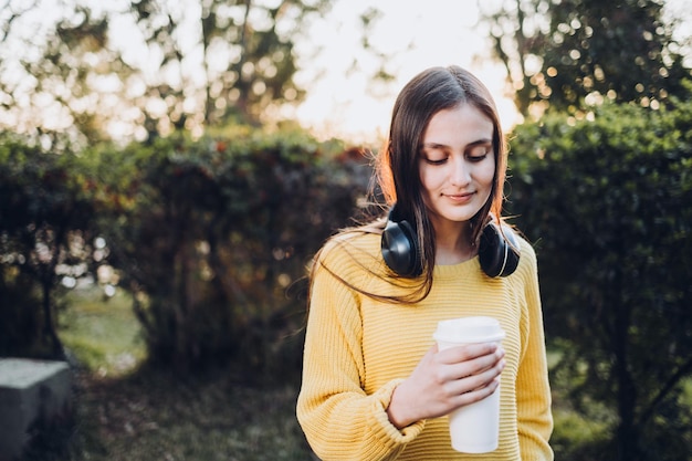 Chillin teenage girl wearing yellow sweater, headphones and holding coffee cup at sunset in the park