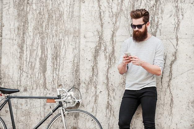 Chilling after good ride. handsome young bearded man holding\
mobile phone while standing near his bicycle against the concrete\
wall