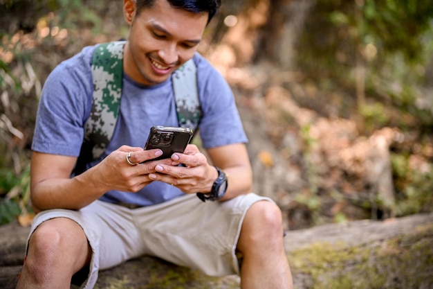 森の中の木の丸太に座りながら、スマートフォンを使って身も凍るようなアジア人の男性トレッカー