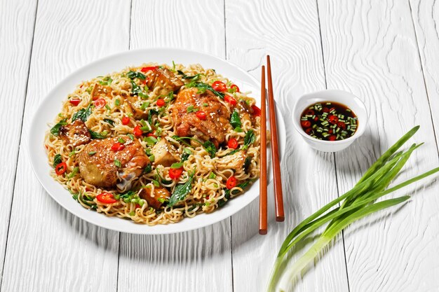 Chinese cuisine: wok noodles with sweet and sour chicken\
thighs, baked tofu, wilted kale, red chili, green onion, on a white\
plate with chopsticks on a white wooden background, top view,\
close-up