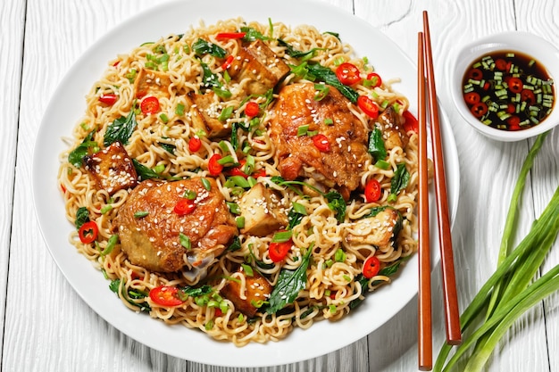 Chinese cuisine: wok noodles with sweet and sour chicken\
thighs, baked tofu, wilted kale, red chili, green onion, on a white\
plate with chopsticks on a white wooden background, top view,\
close-up