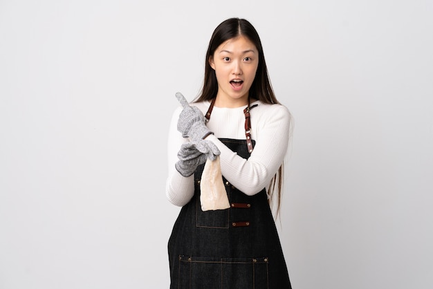 Chinese fishmonger wearing an apron and holding a raw fish over isolated white background surprised and pointing side