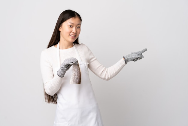 Chinese fishmonger wearing an apron and holding a raw fish over isolated white wall pointing finger to the side and presenting a product