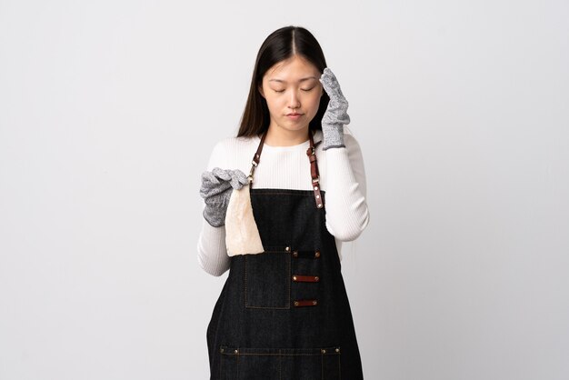 Chinese fishmonger wearing an apron and holding a raw fish over isolated white wall with headache