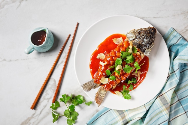 Chinese food: carp squirrel or songshu yu under sweet and sour sauce and fresh cilantro served on a white plate on a white marble stone background, top view, close-up