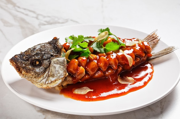 Chinese food: carp squirrel or songshu yu under sweet and sour sauce and fresh cilantro served on a white plate on a white marble stone background, top view, close-up