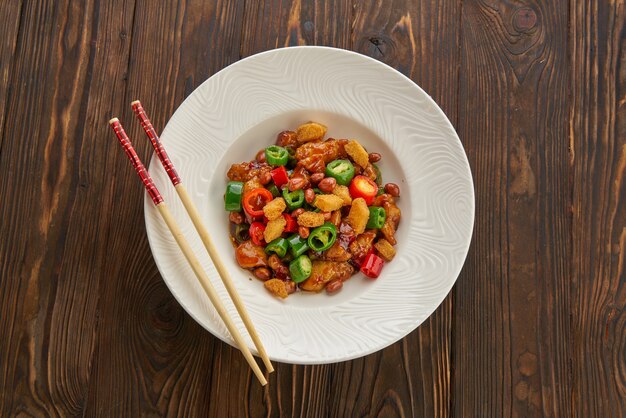 Chinese food, kung pao chicken in white plate with chopsticks on wooden table, top view and copy space