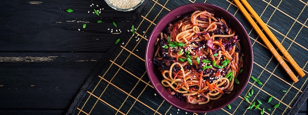 Chinese food. Vegan stir fry noodles with red cabbage and carrot in a bowl on a black wooden background. Asian cuisine meal. Banner. Top view