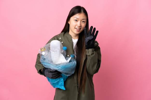 Ragazza cinese che tiene un sacchetto pieno di bottiglie di plastica per riciclare su felice isolato rosa e contando quattro con le dita