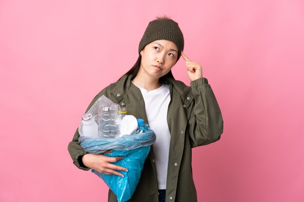 고립 된 분홍색을 통해 재활용 플라스틱 병으로 가득 찬 가방을 들고 중국 소녀는 머리에 손가락을 넣어 광기의 제스처를 만드는