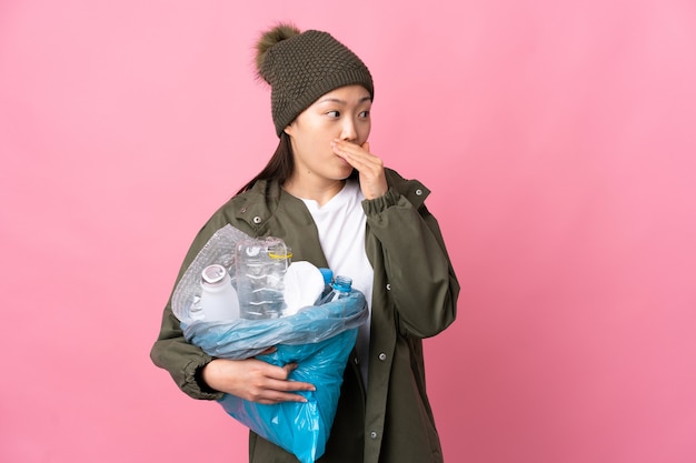 Ragazza cinese che tiene una borsa piena di bottiglie di plastica da riciclare sopra la parete rosa isolata che fa il gesto di sorpresa mentre guarda al lato