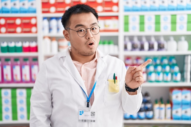 Chinese jonge man aan het werk bij apotheek drogisterij verbaasd wijzend met de vinger naar de zijkant open mond verbaasde uitdrukking