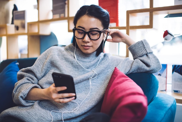 Chinese millennial female reading book online on smartphone\
using earphones for audio