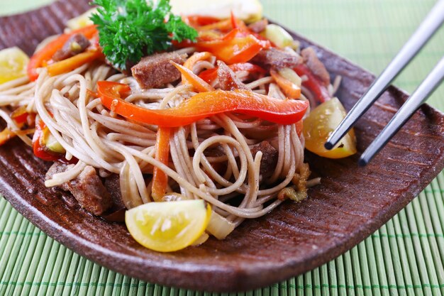 Chinese noodles with vegetables and roasted meat on plate on bamboo mat background