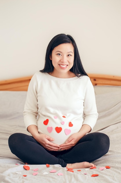 사진 침대에 앉아서 배를 만지는 중국 임신한 여성 임신한 배는 은 심장으로 여 있습니다.