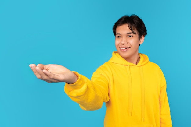 Chinese teen guy stretching hand showing something over blue background