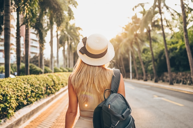 Chinese tourist walking in city streets on Asia vacation tourism. Caucasian beautiful woman in hat with backpack travel lifestyle concept. Walking street, green palms and highway in Sanya, Hainan.