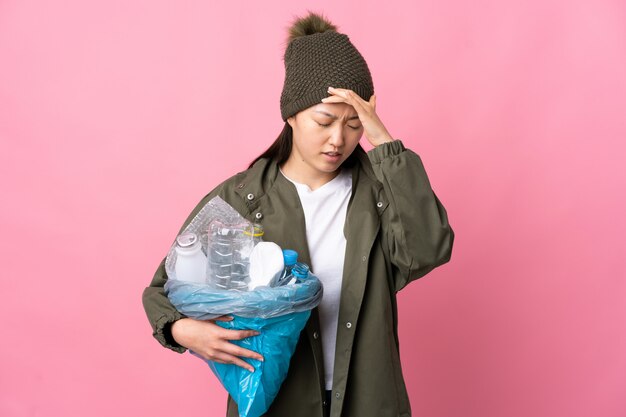 Chinese woman holding a bag full of plastic bottles to recycle over isolated pink wall with headache