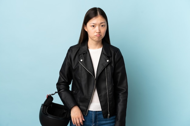 Chinese woman holding a motorcycle helmet over isolated blue background sad