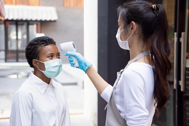 Foto personale di vendita al dettaglio di donna cinese o negoziante di alimentari che controlla la febbre del cliente africano nero tramite un dispositivo di scansione termica portatile