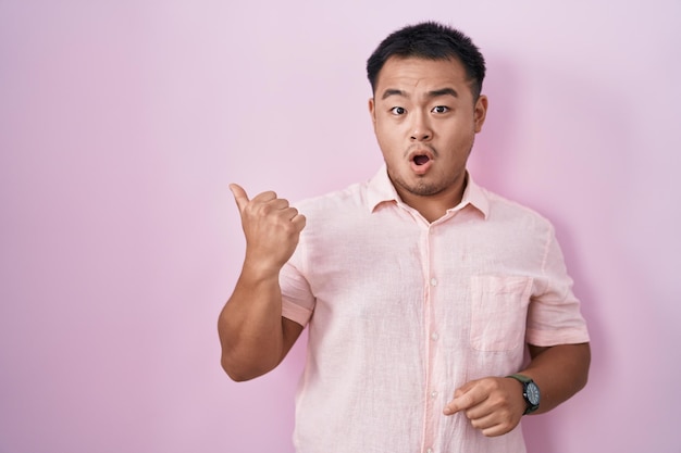 Chinese young man standing over pink background surprised pointing with hand finger to the side open mouth amazed expression