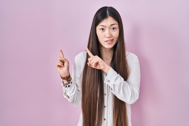 분홍색 배경 위에 서 있는 중국 젊은 여성은 두 손을 걱정하고 놀란 표정으로 걱정과 긴장을 옆으로 향하고 있습니다.