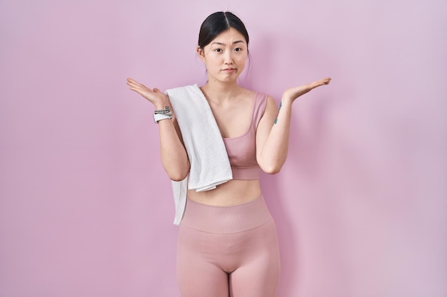 Chinese young woman wearing sportswear and towel clueless and confused expression with arms and hands raised. doubt concept.