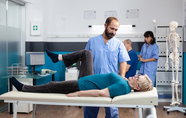 Chiropractor giving assistance to senior patient with knee\
injury for physical recovery. woman doing leg exercise with help\
from medical assistant for osteopathic remedy and treatment.