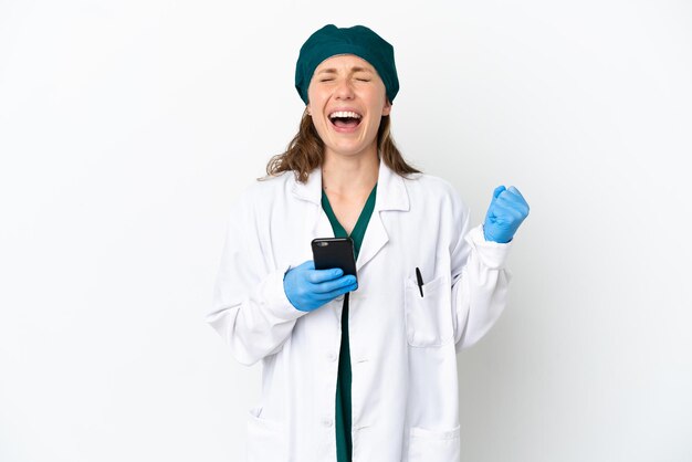 Chirurg blanke vrouw in groen uniform geïsoleerd op een witte achtergrond met telefoon in overwinningspositie