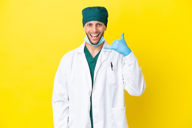 Chirurg blonde man in groen uniform geïsoleerd op gele achtergrond telefoon gebaar maken Bel me terug teken