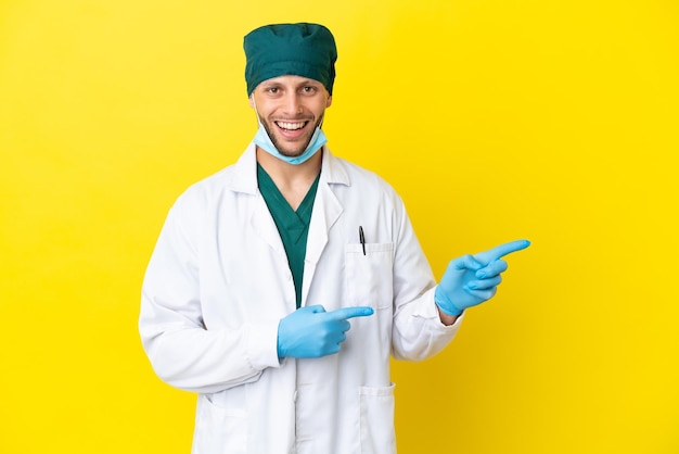 Chirurg blonde man in groen uniform geïsoleerd op gele achtergrond verrast en wijzende kant