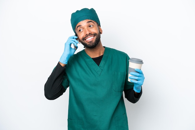 Chirurg Braziliaanse man in groen uniform geïsoleerd op een witte achtergrond met koffie om mee te nemen en een mobiel