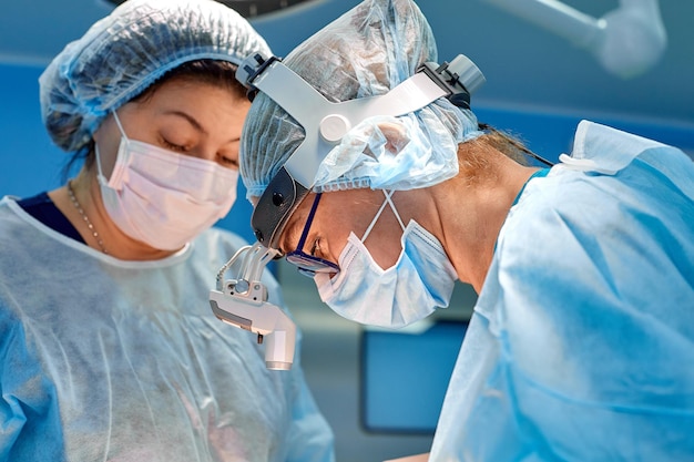 Chirurg die cosmetische chirurgie uitvoert in de operatiekamer van het ziekenhuis Chirurg met masker die loepen draagt tijdens medische procedure Borstvergroting vergrotingsverbetering Blauwe achtergrond blauwe filter