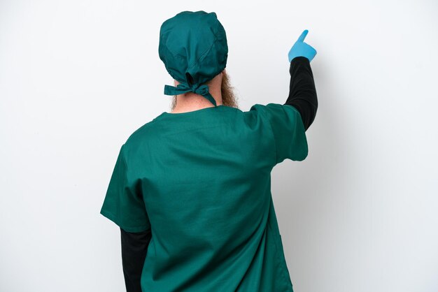Chirurg roodharige man in groen uniform geïsoleerd op een witte achtergrond terug te wijzen met de wijsvinger
