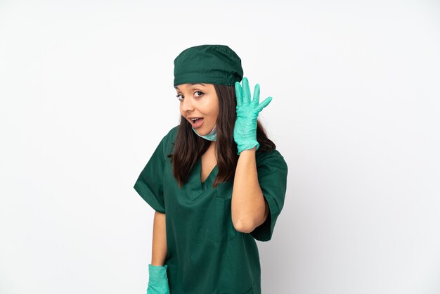 Chirurgenvrouw in groen uniform geïsoleerd op witte muur die aan iets luistert door hand op het oor te zetten