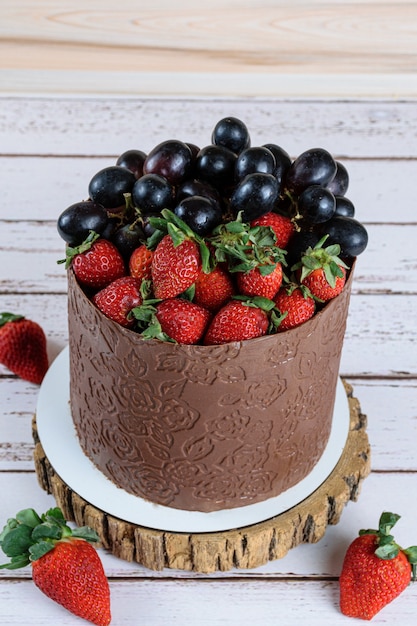 Chocolade aangeklede cake, versierd met druiven en aardbeien, op een witte houten tafel (verticale foto).