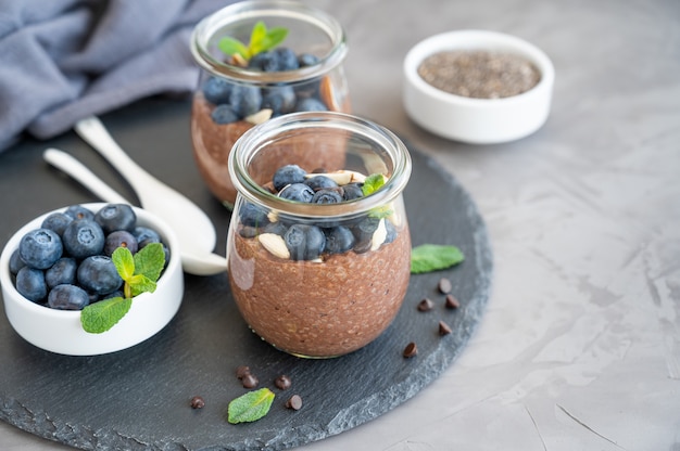 Chocolade chia pudding met bosbessen, amandelen en munt bovenop in een glazen pot op een grijze betonnen ondergrond. Gezond eten. Ruimte kopiëren.