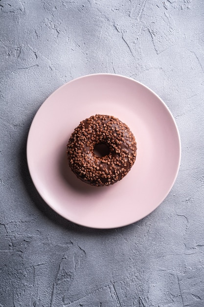Chocolade donut met hagelslag op roze plaat, zoet geglazuurd dessertvoedsel op betonnen getextureerde tafel, bovenaanzicht