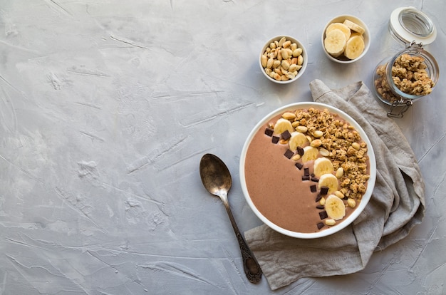 Chocolade smoothie kom met bananen, muesli en pinda's op lichtgrijze betonnen ondergrond. gezond vegetarisch ontbijt. bovenaanzicht met ruimte voor tekst.