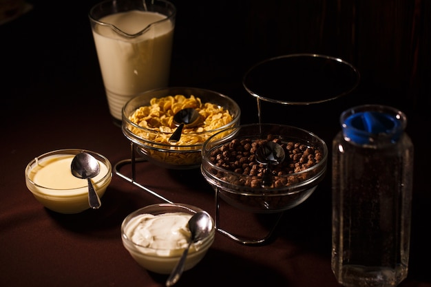 Chocoladeballetjes in een kom en cornflakes met een kan melk en water op tafel en een kom zure room en gecondenseerde melk. Heerlijk en gezond ontbijt